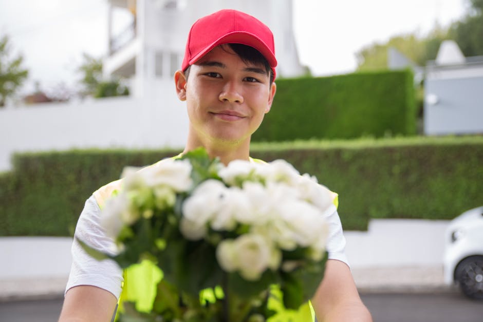 A delivery person wearing a red cap and a reflective yellow vest is extending a bouquet of fresh white flowers, including roses and greenery, towards the camera on a residential street. The bouquet is