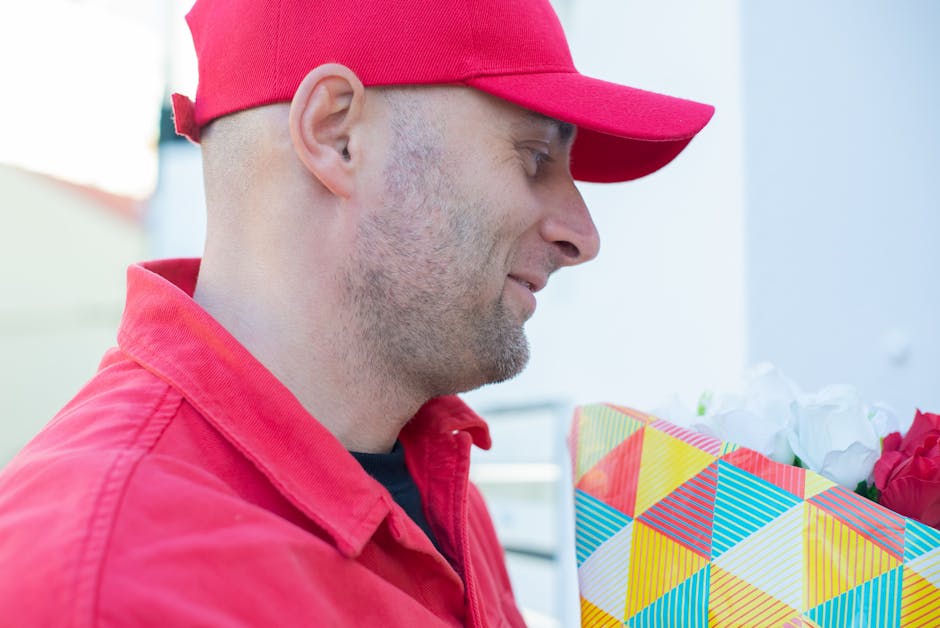 A man dressed in a red baseball cap and red jacket is holding a floral gift wrapped in a colorful paper with geometric patterns, featuring yellow, blue, orange, and white triangles. The bouquet contai