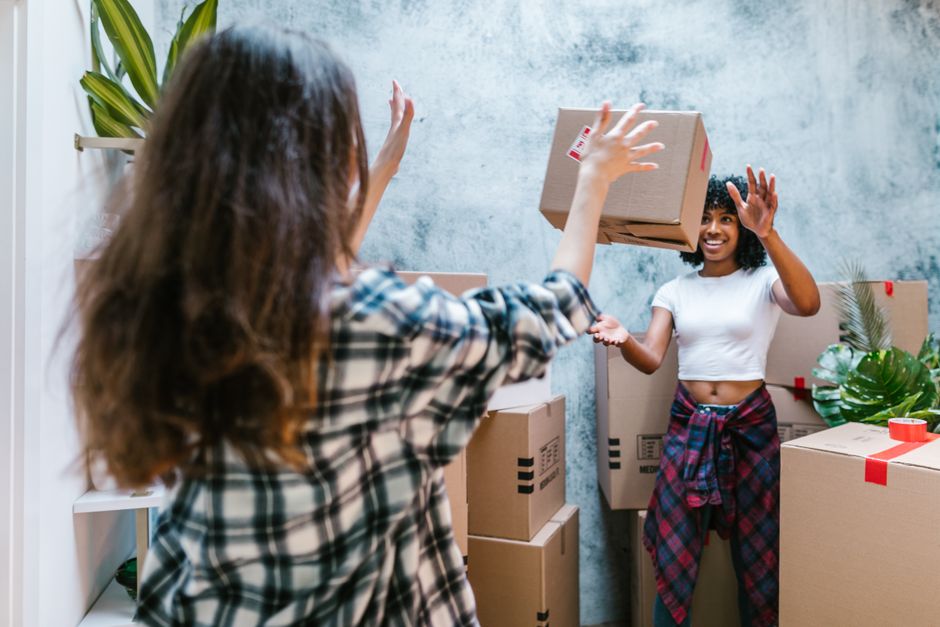 A woman with long brown hair, wearing a plaid shirt, is extending her hands to pass a small cardboard box to a smiling woman with curly dark hair, dressed in a white crop top and plaid skirt, in a roo