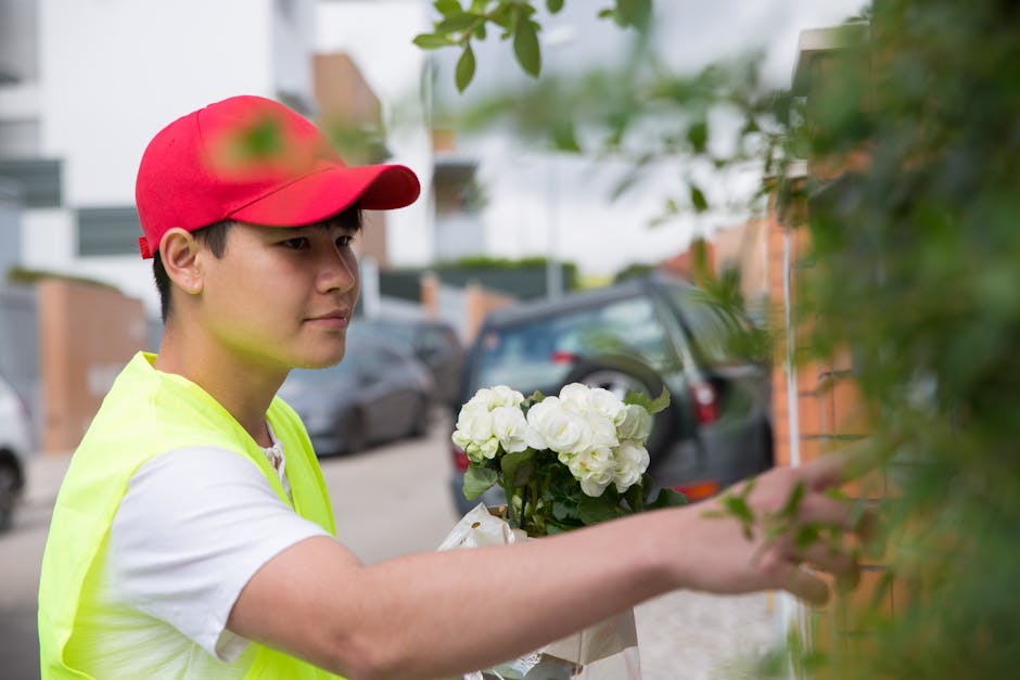 A young delivery person wearing a red cap and a high-visibility yellow vest arranges a bouquet of fresh white hydrangeas wrapped in white paper, with some greenery, against a brick wall outdoors durin