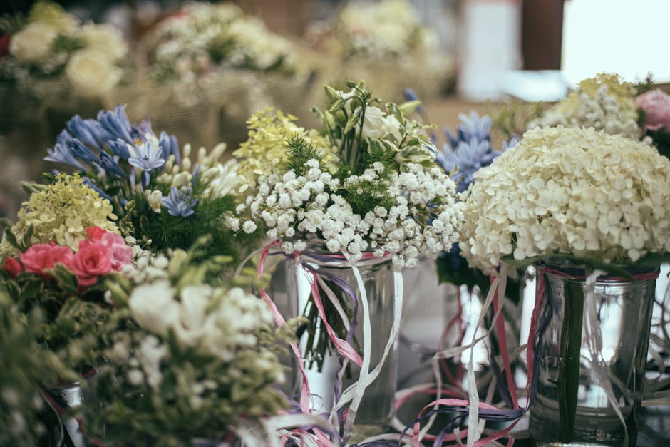 A floral arrangement featuring white hydrangeas, delicate white baby's breath, and soft pink roses, displayed in clear glass vases wrapped with pink and white ribbon. The bouquets are arranged on a ta
