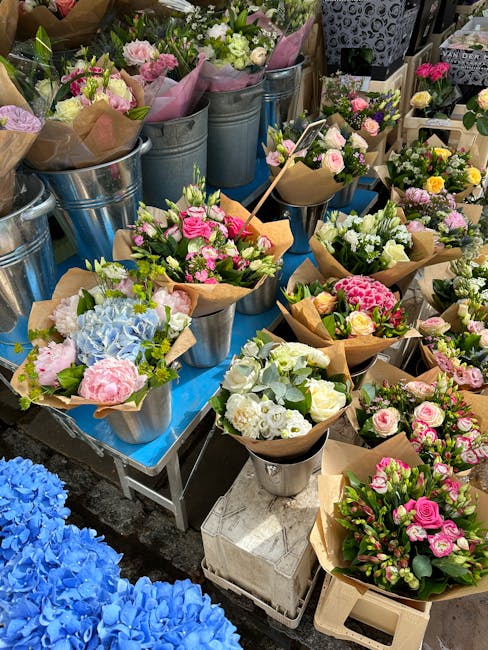 An outdoor display of freshly arranged floral bouquets featuring pastel pink roses, white and green hydrangeas, and delicate filler flowers wrapped in brown kraft paper, placed in silver and ceramic b