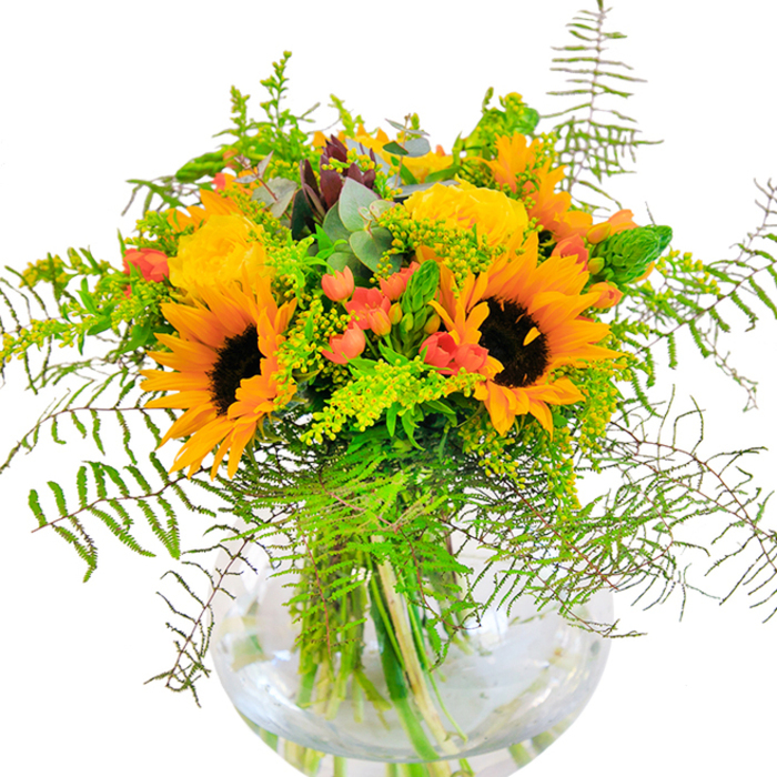 Upon first encountering this bouquet, one is greeted with a wave of golden radiance-a jubilant gathering of sunflowers and yellow garden roses, meticulously arranged by a knowledgeable New Southgate florist with an eye for seasonal celebration. The sunflowers, with their broad, dark-centered faces, seem to reach out toward the viewer, while the buttery garden roses unfurl in sumptuous spirals, their texture soft and plush beneath the light. Glossy red berries of Hypericum nestle amongst layers of varied greenery: the fine lace of ferns, the soft haze of light-green filler, and elongated sprays of goldenrod brimming with tiny yellow blooms, each layer enhancing the arrangement's depth and natural movement. Eucalyptus leaves, with their muted blue-green tones, peek through the display, adding a subtle rustic fragrance like a gentle breeze along Brunswick Park Road. The bouquet rests in a perfectly round, clear glass vase, enchanting in its clarity and providing a window to the vibrant gathering of stems below-a vignette of life and growth reminiscent of New Southgate's own blend of energy and tranquility. Whether gifted for meaningful milestones or placed as a bright centrepiece in a family home, this bouquet exudes both joy and elegance, infusing every New Southgate occasion with colour, freshness, and heartfelt beauty.